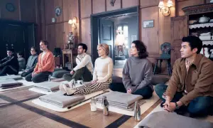 Un gruppo di nove perfetti sconosciuti medita in una stanza di legno, ognuno su un tappetino con coperte piegate.