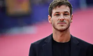 Ritratto di Gaspard Ulliel, attore morto oggi, con un blazer scuro su sfondo rosa sfocato.