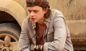 Dane DeHaan, con bandana e collana, pensieroso accanto a un veicolo fangoso in un film.