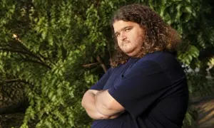 Jorge Garcia, noto per i suoi ruoli in film e programmi TV, con i suoi iconici capelli ricci e una maglietta blu, posa con le braccia conserte.