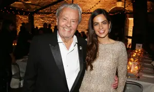 Alain Delon oggi, sorridente con una giovane donna elegante, durante un evento serale illuminato da lucine.