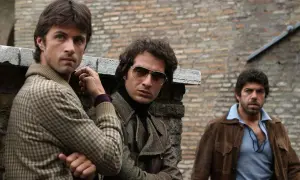 Il cast di Romanzo Criminale - La Serie: tre uomini con abiti anni '70, uno con occhiali da sole, appoggiati a un muro di mattoni.
