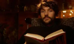 Un giovane uomo con barba e capelli castani, parte del cast di Merlin, legge un libro aperto seduto su una poltrona.