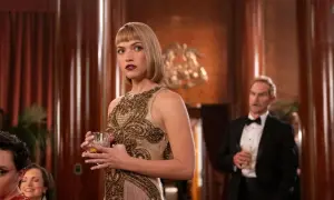 La donna con abito dorato e frange tiene un bicchiere. Sullo sfondo, un uomo in smoking e altri dettagli suggeriscono un'atmosfera da "cast di morte".