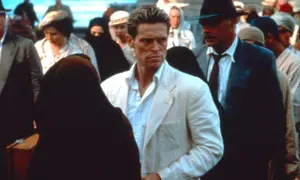 Willem Dafoe in un film, con un completo bianco, circondato da persone in abiti tradizionali e un uomo con cappello.