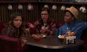 Benjamin Flores Jr. sorride con un cappello e una collana d'oro, mentre due ragazze discutono al tavolo di un diner.