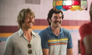 Due uomini sorridono, uno con capelli biondi e baffi, l'altro con capelli scuri e baffi. Scena da un film di Glen Powell.