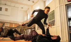 Scena di combattimento intensa, una donna in tuta nera salta su un uomo a terra. Un altro uomo è a terra in background. Un film di 60 minuti pieno d'azione.