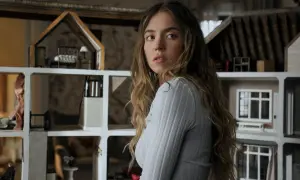 Una giovane donna con lunghi capelli castani guarda intensamente, circondata da case delle bambole. Sembra una di famiglia, forse la housemaid che osserva la vita degli altri.