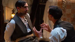 Un uomo legato con una corda, con macchie di sangue sulla camicia, ascolta attentamente un altro uomo che gesticola. La promessa anticipazioni.