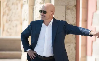 Un uomo calvo con occhiali da sole, giacca blu e camicia bianca, pronto per un'indagine in una delle serie tv poliziesche più avvincenti.
