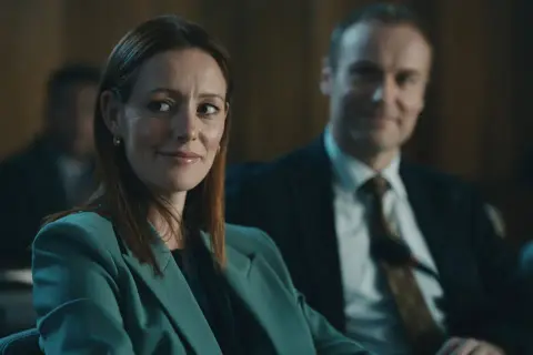 Donna con tailleur verde acqua sorride, mentre un uomo in giacca e cravatta la osserva. Forse stanno discutendo del codice da un miliardo di dollari.