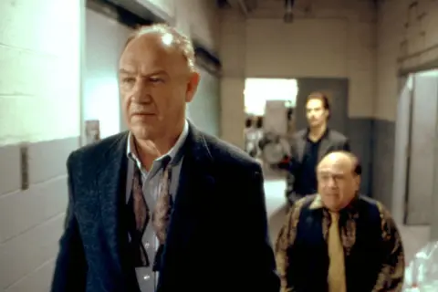 Gene Hackman in un film, cammina con altri due uomini in un corridoio.