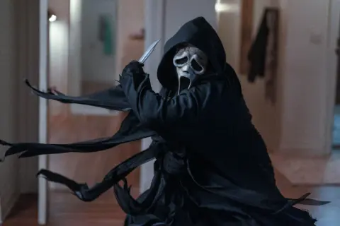 Ghostface, icona del film di Scream, brandisce un coltello con la sua maschera spettrale e il mantello svolazzante.