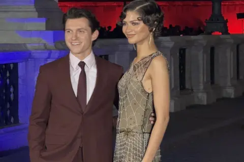 Tom Holland e Zendaya, una coppia iconica, sorridono alla telecamera. Lui indossa un completo bordeaux, lei un abito scintillante.