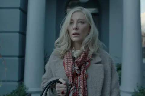 Donna bionda con foulard rosso e cappotto beige, espressione preoccupata. Un'immagine che anticipa un disclaimer serie tv.