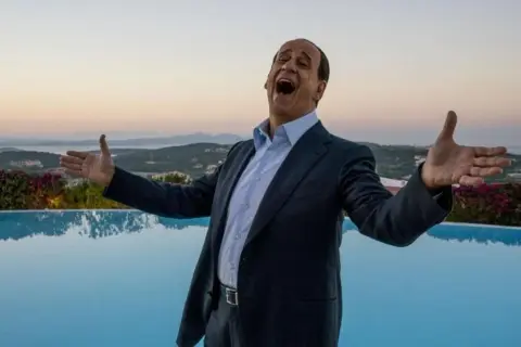 Toni Servillo nei panni di Berlusconi, con le braccia aperte e la bocca spalancata, celebra un momento di gioia davanti a una piscina e al mare al tramonto.