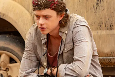 Dane DeHaan, con bandana e collana, pensieroso accanto a un veicolo fangoso in un film.