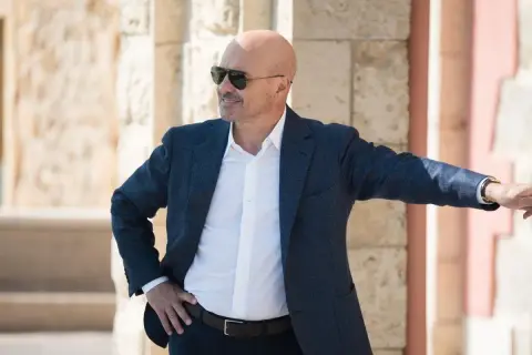 Un uomo calvo con occhiali da sole, giacca blu e camicia bianca, pronto per un'indagine in una delle serie tv poliziesche più avvincenti.