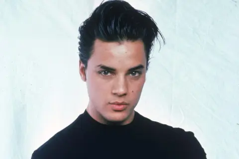 Ritratto di Nick Kamen, un giovane uomo con capelli scuri pettinati all'indietro e un maglione nero. La sua espressione è seria, mentre si riflette sulla sua **malattia**.