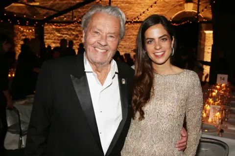 Alain Delon oggi, sorridente con una giovane donna elegante, durante un evento serale illuminato da lucine.