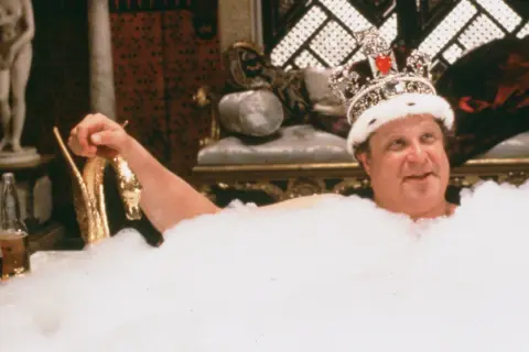 Un uomo con una corona scintillante, immerso in una vasca piena di schiuma, sorride. Sembra che sua maestà viene da Las Vegas, pronto per un bagno regale.