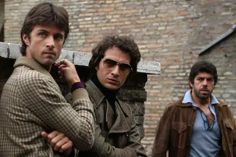 Il cast di Romanzo Criminale - La Serie: tre uomini con abiti anni '70, uno con occhiali da sole, appoggiati a un muro di mattoni.