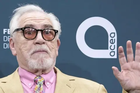 Brian Cox, noto per i suoi film e programmi TV, con occhiali e cravatta colorata.