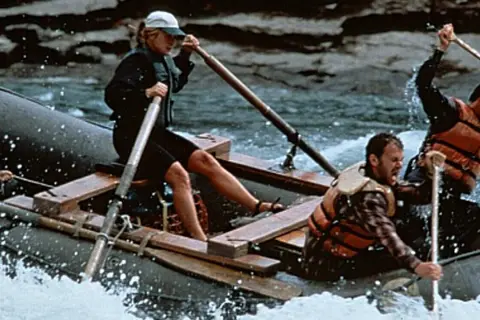 Il cast di The River Wild affronta rapide impetuose su un gommone, remando con forza per sopravvivere.