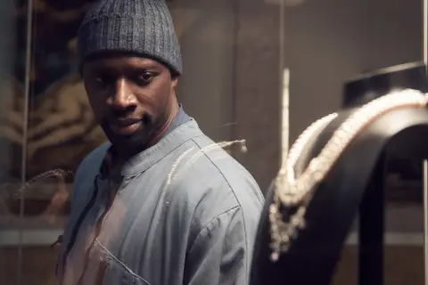 Omar Sy, protagonista di film e programmi TV, osserva con astuzia una collana di perle in una vetrina.