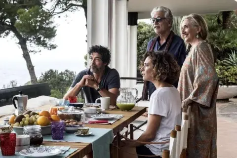 Una famiglia riunita per una colazione all'aperto, tra risate e sguardi pensierosi. Un momento di gioia e riflessione, una vera croce e delizia.