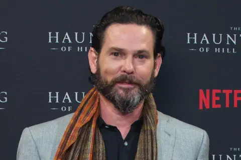Henry Thomas, noto per i suoi film e programmi TV, sorride con una sciarpa colorata.