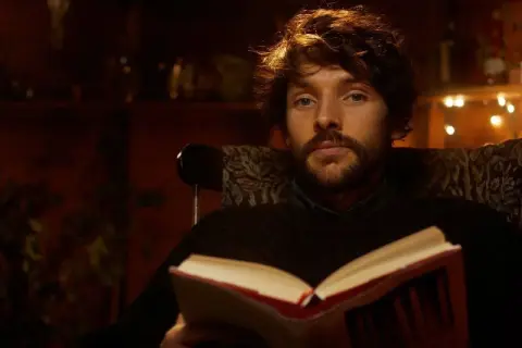 Un giovane uomo con barba e capelli castani, parte del cast di Merlin, legge un libro aperto seduto su una poltrona.