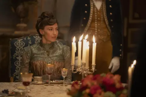 Una donna elegante, in abito d'epoca, siede a tavola durante un sontuoso banchetto, un'immagine che evoca lo sfarzo e le tensioni sociali della Gilded Age.
