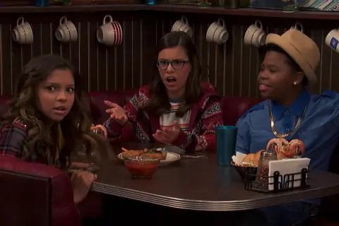 Benjamin Flores Jr. sorride con un cappello e una collana d'oro, mentre due ragazze discutono al tavolo di un diner.