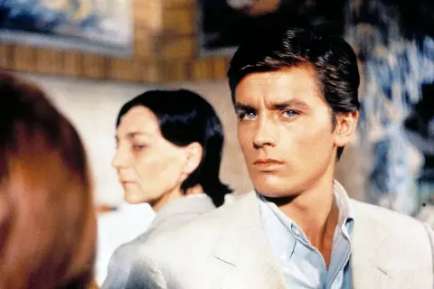 Alain Delon, icona di stile, in una scena che evoca i suoi film più belli. Sguardo intenso, fascino intramontabile.