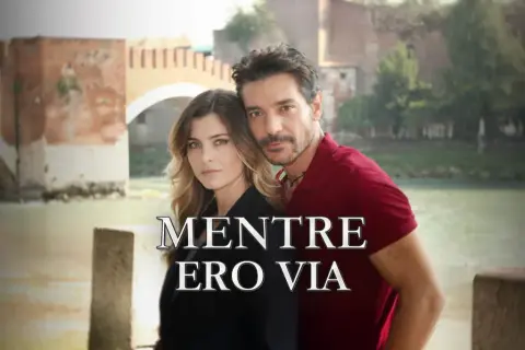 Coppia abbracciata con un ponte sullo sfondo. Il titolo "Mentre ero via" suggerisce una storia di assenza e ritorno.