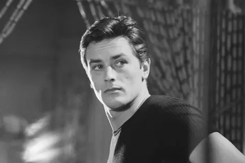Alain Delon da ragazzo, con sguardo intenso e capelli pettinati all'indietro, indossa una maglia a coste.
