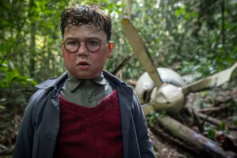 Ragazzo con occhiali e segni sul viso, in una foresta, vicino a un relitto d'aereo. Scena da "Il signore delle mosche serie tv".