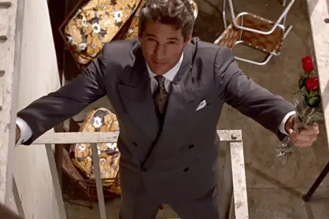 Richard Gere, iconico in "American Gigolo film", con un mazzo di rose rosse, elegante in abito grigio.