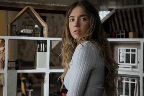 Una giovane donna con lunghi capelli castani guarda intensamente, circondata da case delle bambole. Sembra una di famiglia, forse la housemaid che osserva la vita degli altri.