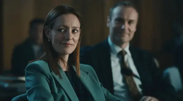 Donna con tailleur verde acqua sorride, mentre un uomo in giacca e cravatta la osserva. Forse stanno discutendo del codice da un miliardo di dollari.