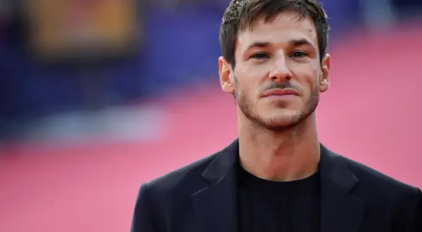 Ritratto di Gaspard Ulliel, attore morto oggi, con un blazer scuro su sfondo rosa sfocato.