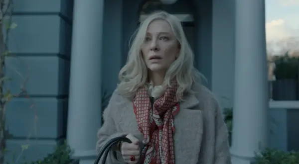 Donna bionda con foulard rosso e cappotto beige, espressione preoccupata. Un'immagine che anticipa un disclaimer serie tv.