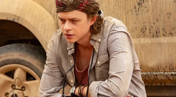 Dane DeHaan, con bandana e collana, pensieroso accanto a un veicolo fangoso in un film.