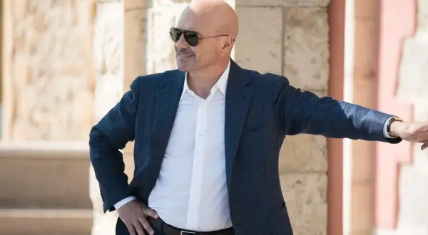 Un uomo calvo con occhiali da sole, giacca blu e camicia bianca, pronto per un'indagine in una delle serie tv poliziesche più avvincenti.