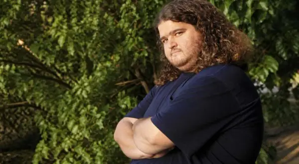 Jorge Garcia, noto per i suoi ruoli in film e programmi TV, con i suoi iconici capelli ricci e una maglietta blu, posa con le braccia conserte.