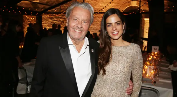 Alain Delon oggi, sorridente con una giovane donna elegante, durante un evento serale illuminato da lucine.