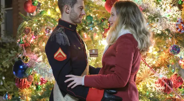 Un uomo in uniforme militare, un principe sotto l'albero di Natale, abbraccia una donna con un cappotto rosso. Le luci scintillanti dell'albero creano un'atmosfera magica.