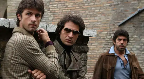 Il cast di Romanzo Criminale - La Serie: tre uomini con abiti anni '70, uno con occhiali da sole, appoggiati a un muro di mattoni.